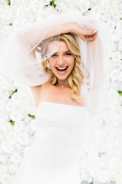 cheerful young bride holding bridal veil in raised hands on white floral background - Фото, изображение
