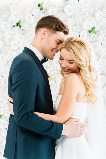 happy beautiful bride hugging smiling groom on white floral background - Zdjęcie, obraz