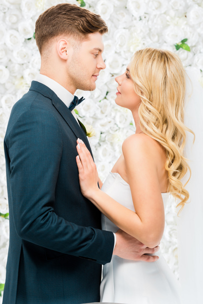 glückliche junge Braut und Bräutigam stehen von Angesicht zu Angesicht auf weißem Blumenhintergrund - Foto, Bild