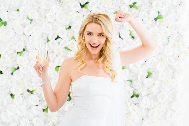 cheerful young bride with glass of champagne looking at camera on white floral background - Φωτογραφία, εικόνα