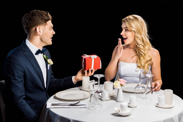 handsome groom presenting red gift box to surprised bride isolated on black - Photo, Image
