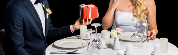 panoramic shot of groom presenting red gift box to bride, while sitting at served table isolated on black - Foto, Imagem