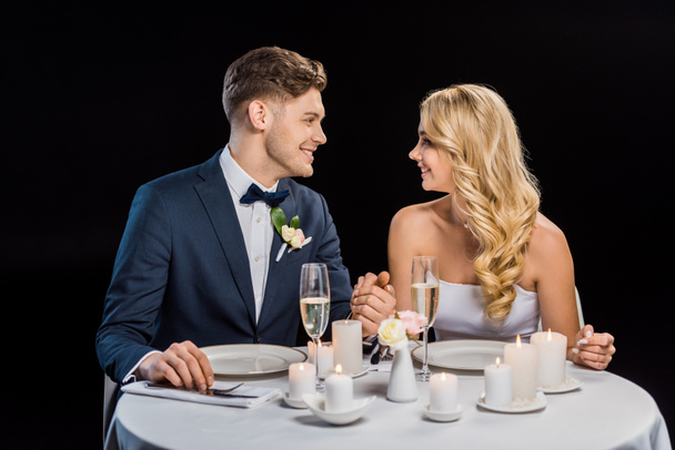 glücklicher junger Bräutigam und Braut Händchen haltend, während sie am gedeckten Tisch isoliert auf schwarz sitzen - Foto, Bild
