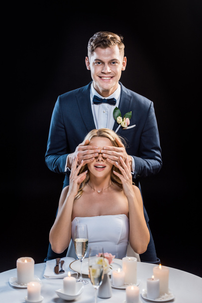 happy groom standing behind smiling bride and closing her eyes with hands isolated on black - Photo, Image