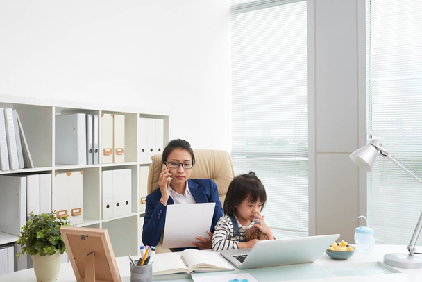 Adult businesswoman having phone call and looking papers with adorable daughter sitting on knees at workplace - Photo, image