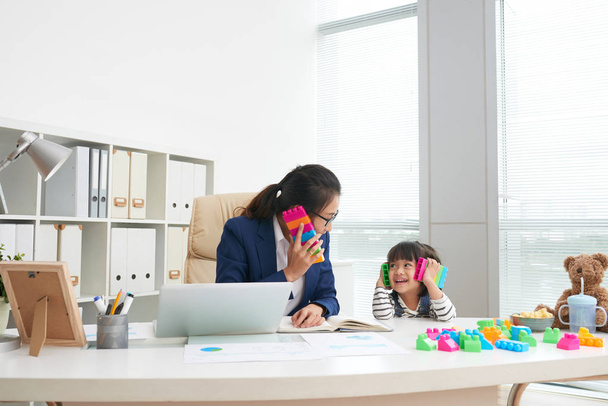 Взрослая азиатская официальная деловая женщина с игривой маленькой девочкой, сидящей за столом в современном офисе, развлекаясь с красочными блоками
 - Фото, изображение