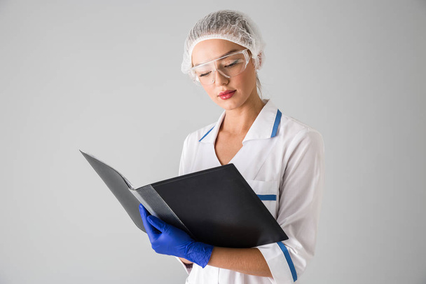 Beautiful young woman cosmetologist doctor isolated over grey wall background holding folder. - Foto, Imagem