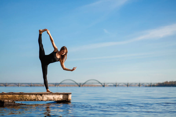 Young woman practicing yoga exercise at quiet wooden pier with city background. Sport and recreation in city rush - Photo, Image