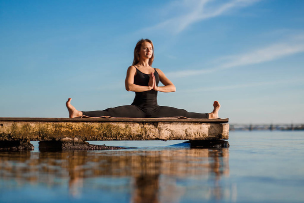 Young woman practicing yoga exercise at quiet wooden pier with city background. Sport and recreation in city rush - 写真・画像