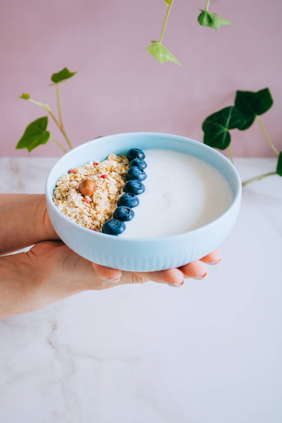 Healthy breakfast bowl with muesli and yogurt in woman hands on pink and marble background. Clean eating, vegetarian, vegan, alkiline diet food concept - Foto, Imagen