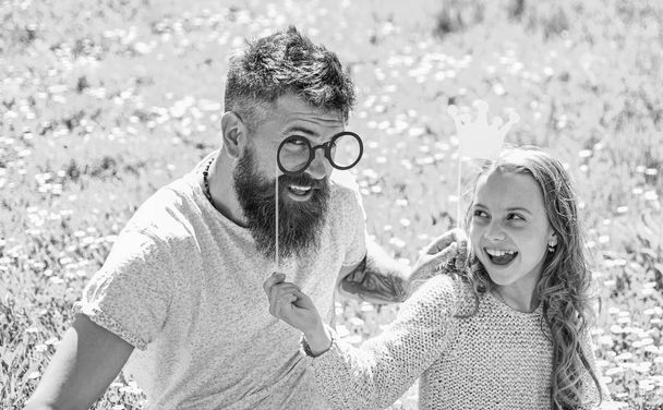 Dad and daughter sits on grass at grassplot, green background. Daddys princess concept. Family spend leisure outdoors. Child and father posing with crown and eyeglasses photo booth attributes - Φωτογραφία, εικόνα
