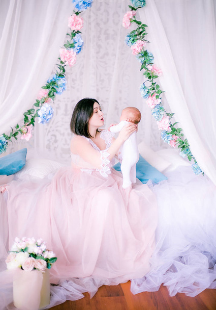 Young mother in a boudoir dress with a baby in her arms in spring flowers by the canopy bed - Photo, image