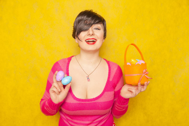 гарна доросла пухка жінка з коротким волоссям, що тримає барвисті святкові яйця та великодній кошик на жовтому твердому студійному фоні
 - Фото, зображення