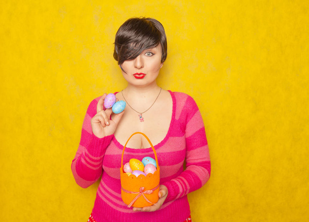 bastante adulto gordita mujer con pelo corto celebración de coloridos huevos de vacaciones y una cesta de Pascua en amarillo sólido fondo del estudio
 - Foto, Imagen