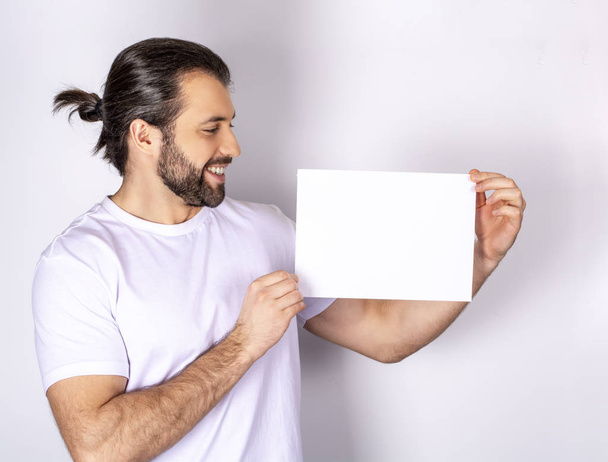 красивый мужчина в белой футболке на белом фоне, показывает белый лист бумаги
 - Фото, изображение