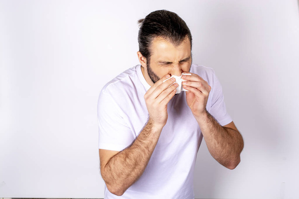 Hombre guapo en una camiseta blanca sobre un fondo blanco, estornuda, coge un resfriado
 - Foto, imagen