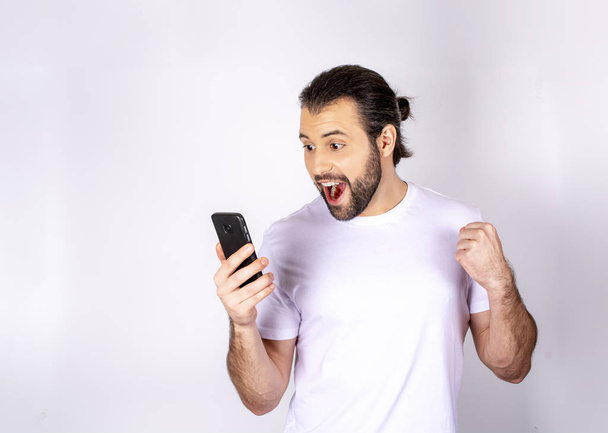 Handsome man in a white T-shirt on a white background, smiling, rejoices, in the hands of a smartphone, writes SMS - Photo, Image