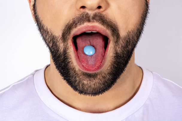 Handsome man in white t-shirt on white background, pill on tongue, close-up - Фото, зображення