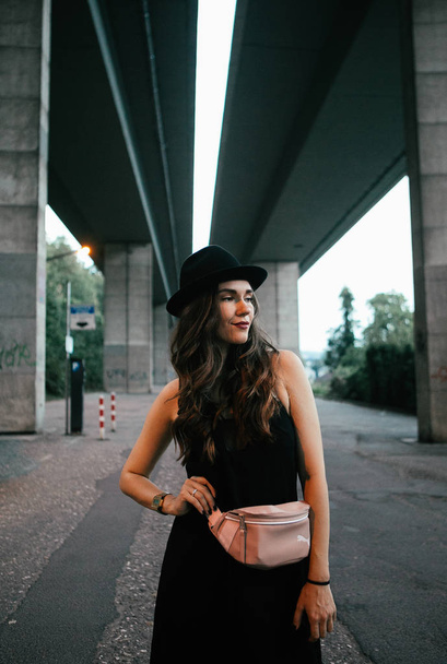Sexy brunetka v černých šatech, klobouk s růžovou kabelku představuje pod kamenný most - Fotografie, Obrázek