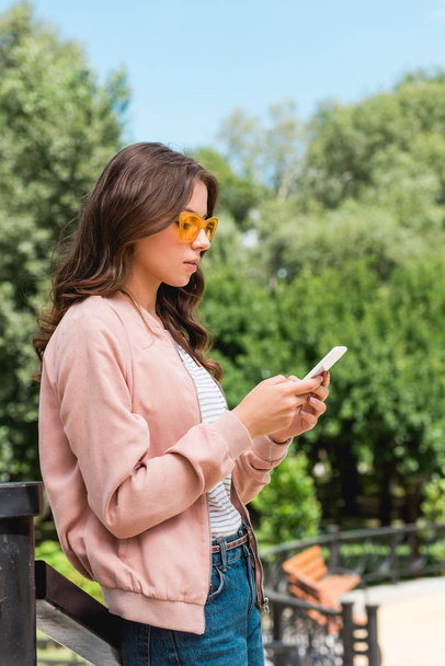 attractive girl in sunglasses using smartphone while standing in park - 写真・画像