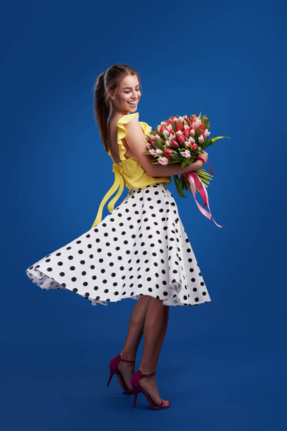 Perfect gift. Wonderful flowers on Womens Day. Tender smiling brunet woman poses with colorful tulips, looks happily, isolated over blue studio background. Space for text - Foto, Imagem