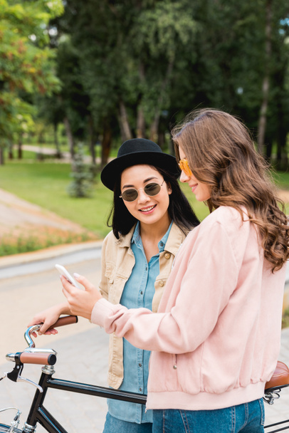 pretty girls in sunglasses smiling while looking at smartphone and standing near bike in park - Photo, image