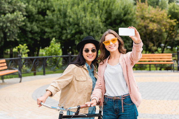 attractive girls in sunglasses smiling while taking selfie in park - 写真・画像