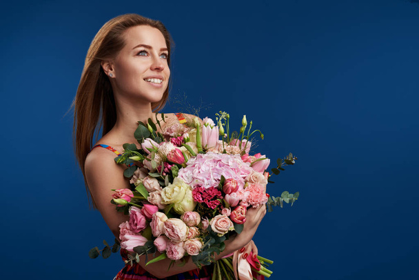 Perfect gift. Wonderful flowers on Womens Day. Tender smiling brunet woman poses with spring bouquet, looks happily, isolated over blue studio background. Space for text - Photo, Image