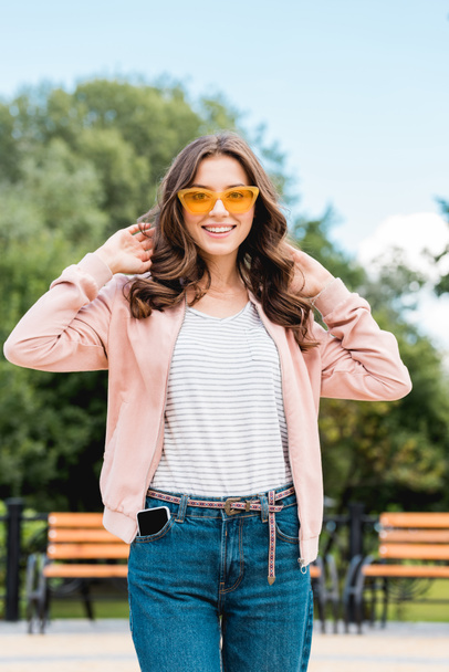 pretty girl in sunglasses smiling while touching hair in park - Foto, Imagem