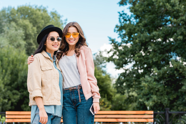 beautiful girls in sunglasses smiling while hugging in park - Фото, изображение