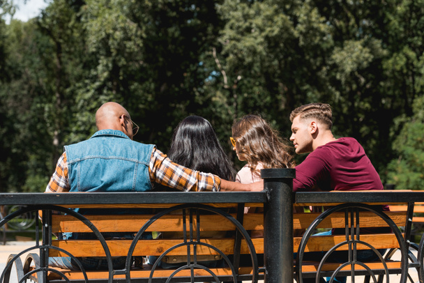 Achteraanzicht van multi-etnische vrienden zitten op bankje in park - Foto, afbeelding