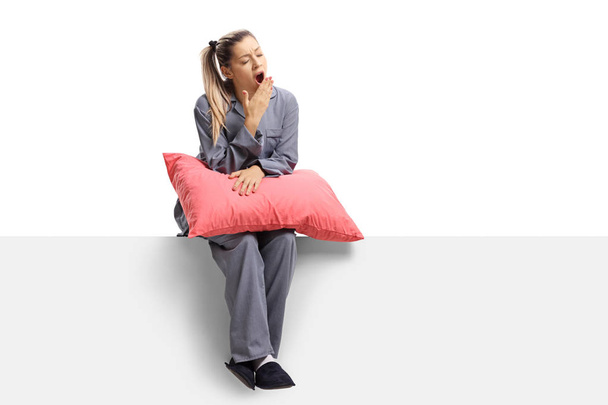 Full length shot of a young sleepy woman sitting on a panel with a pillow and yawning isolated on white background - Photo, Image