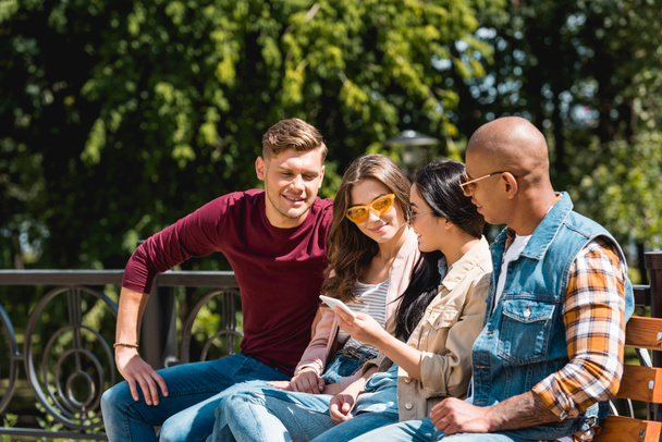cheerful multicultural friends sitting on bench and looking at girl holding smartphone - Photo, Image