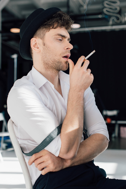 dreamy handsome man in white shirt and black hat smoking cigarette - Foto, afbeelding