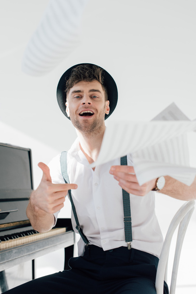 músico feliz en sombrero negro y camisa blanca lanzando hojas de libros de música
  - Foto, Imagen