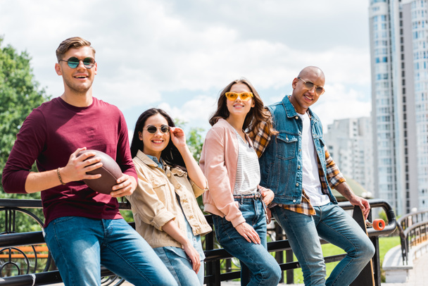 jovens multiculturais felizes em óculos de sol de pé com longboard e futebol americano perto de meninas atraentes
 - Foto, Imagem