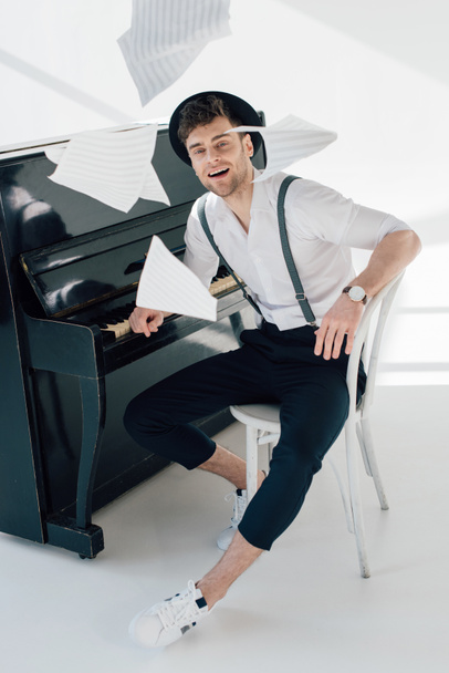 smiling musician in trendy clothing sitting by piano and throwing music book sheets  - Photo, Image