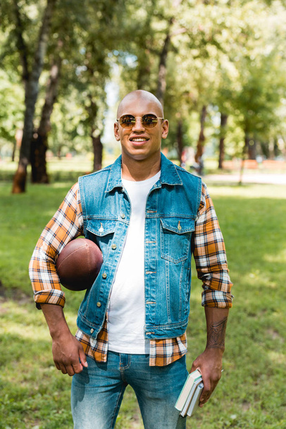 allegro uomo afroamericano in occhiali da sole che tiene libri e football americano
 - Foto, immagini