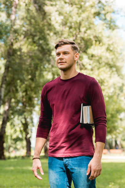 handsome student standing with books and smiling in park - Фото, изображение
