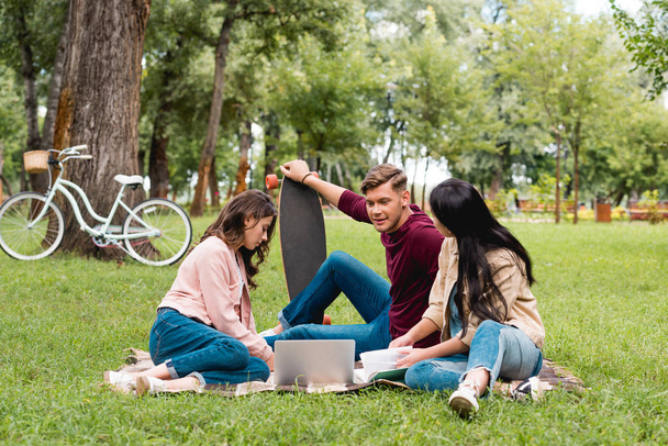 handsome man holding longboard and sitting with girls on blanket near laptop  - Foto, Imagem