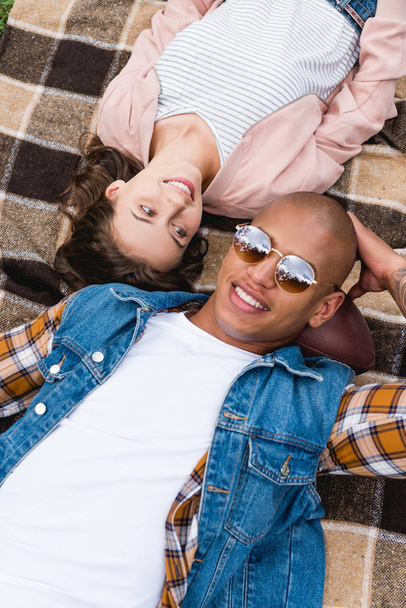 overhead view of cheerful interracial couple lying on checkered blanket - 写真・画像
