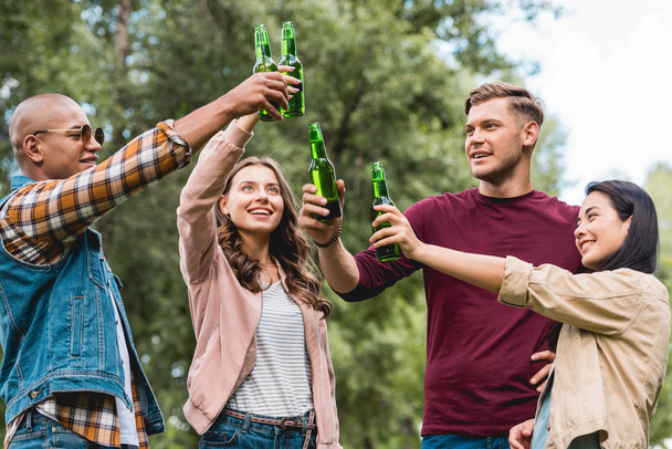 happy multicultural friends clinking bottles with beer while standing in park - Фото, изображение