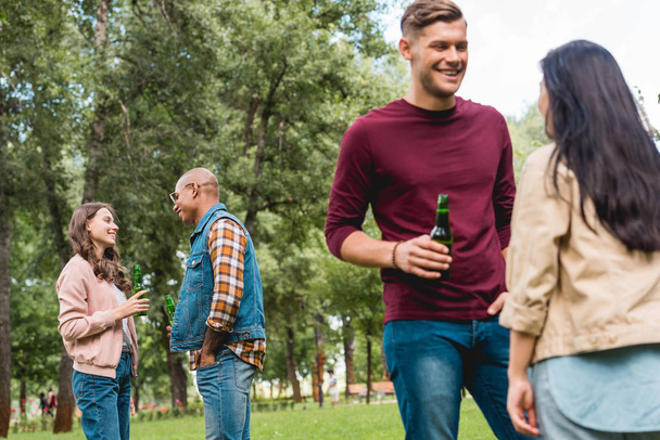 amigos multiculturales felices hablando mientras sostiene botellas con cerveza en el parque
 - Foto, imagen