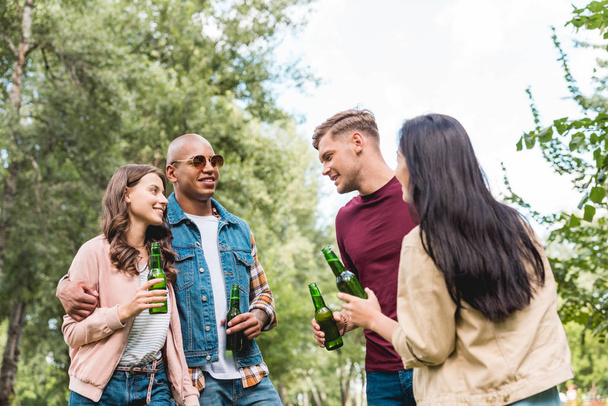 cheerful multicultural friends holding bottles with beer and talking in park - Photo, Image