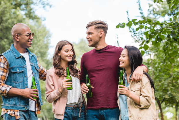 alegre grupo multicultural de amigos sosteniendo botellas con cerveza y hablando en el parque
 - Foto, Imagen