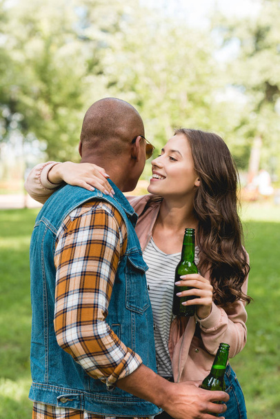 cheerful girl whispering in ear of african american boyfriend while holding bottle  - Foto, immagini