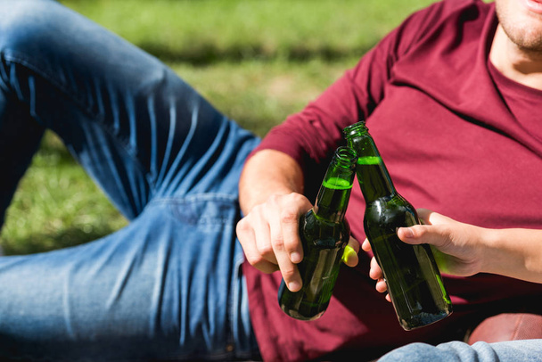 cropped view of couple toasting bottles with beer while lying in park - Valokuva, kuva