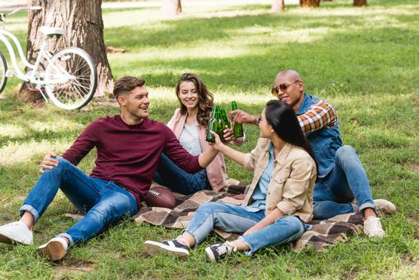 multiethnic group of friends sitting on plaid blanket and clinking bottles in park - Foto, imagen