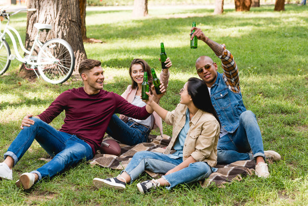 fröhliche multiethnische Gruppe von Freunden, die auf karierter Decke im Park sitzen und Flaschen klirren - Foto, Bild