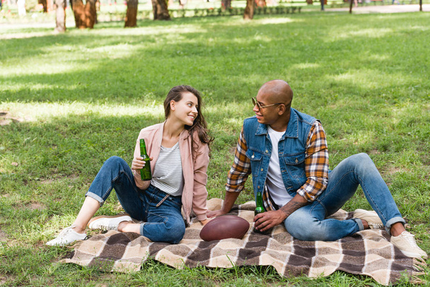 happy interracial couple looking at each other while sitting on blanket with bottles of beer - 写真・画像